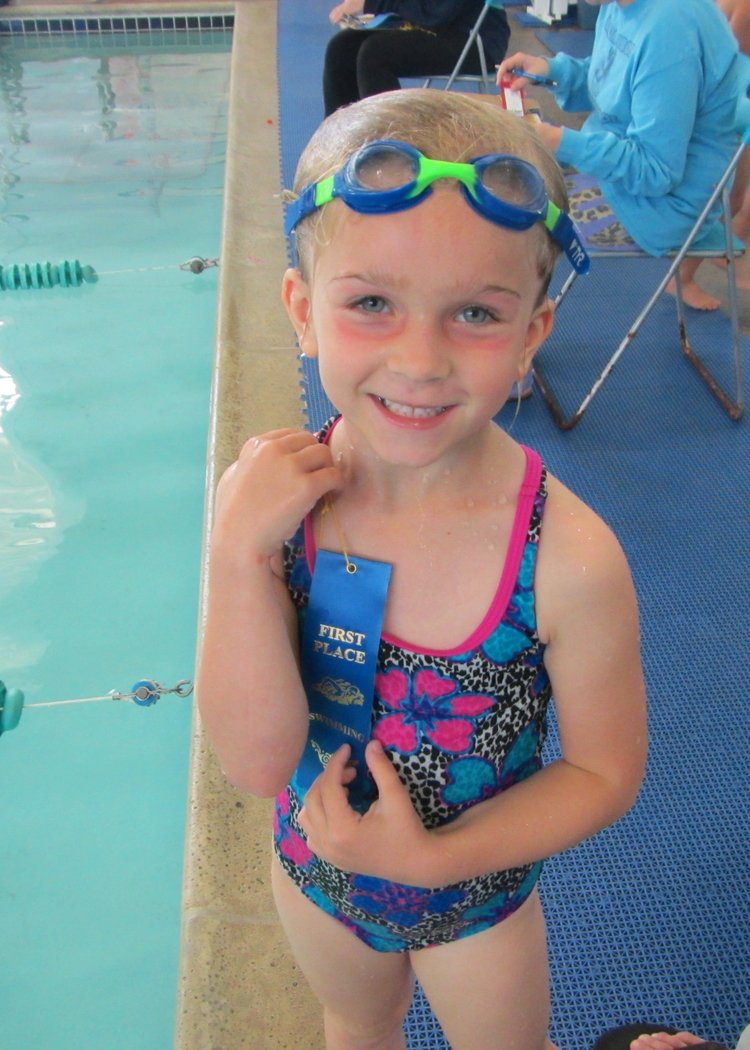 A young girl in a swimsuit and goggles holding a blue ribbon while she stands next to a swimming pool
